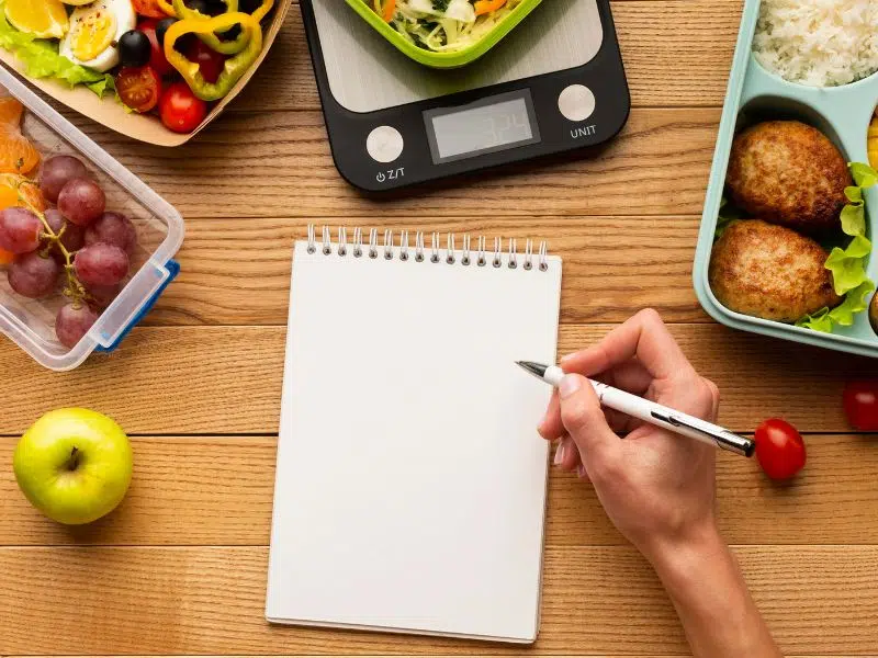 Person writing in a blank notepad surrounded by healthy food and a kitchen scale.